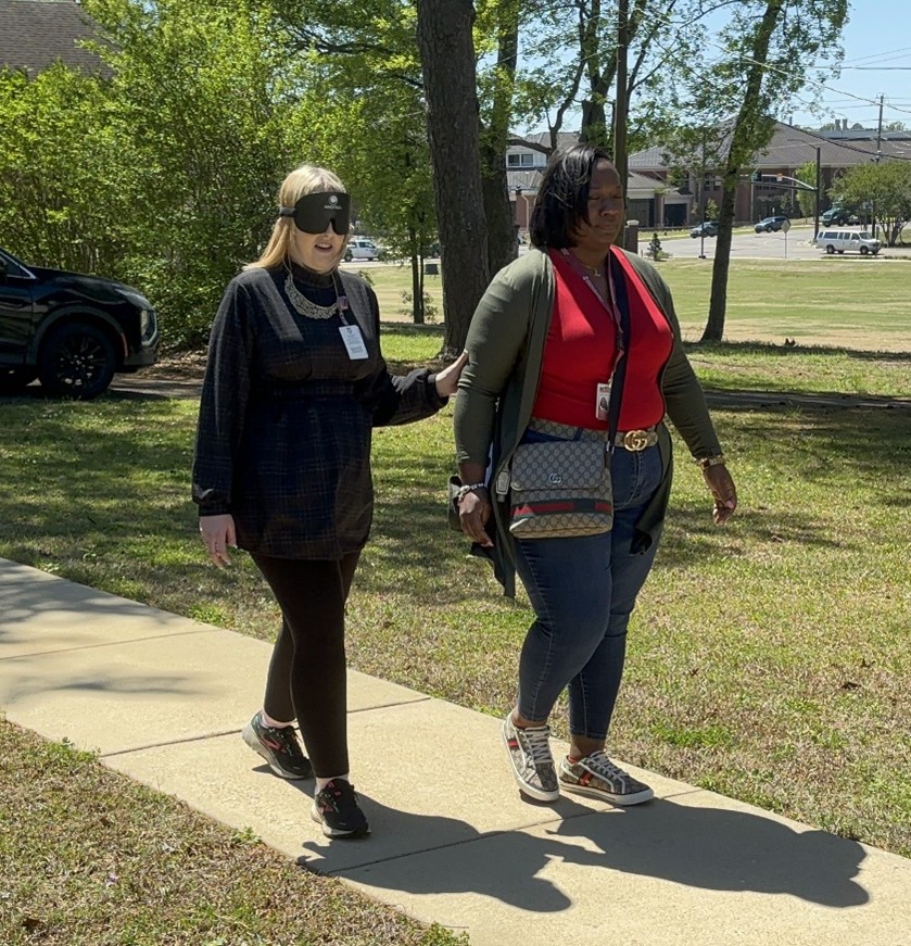 Two women are walking outside on a sunny day, practicing human guide techniques. One woman is wearing a visor and touching the arm of the other woman, who is guiding her.