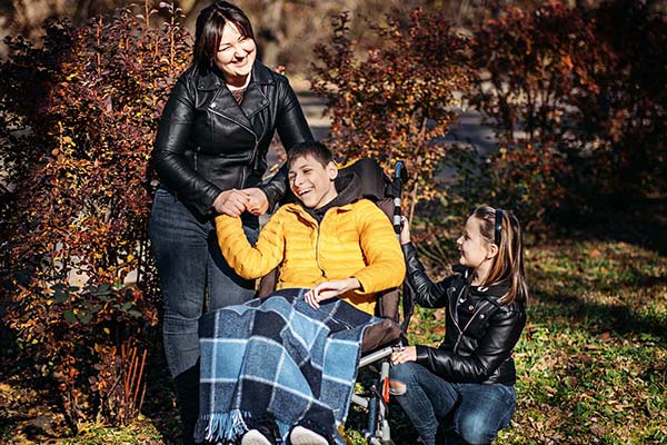 A mother holds her son's hand in a wheelchair as her young daughter kneels beside him.