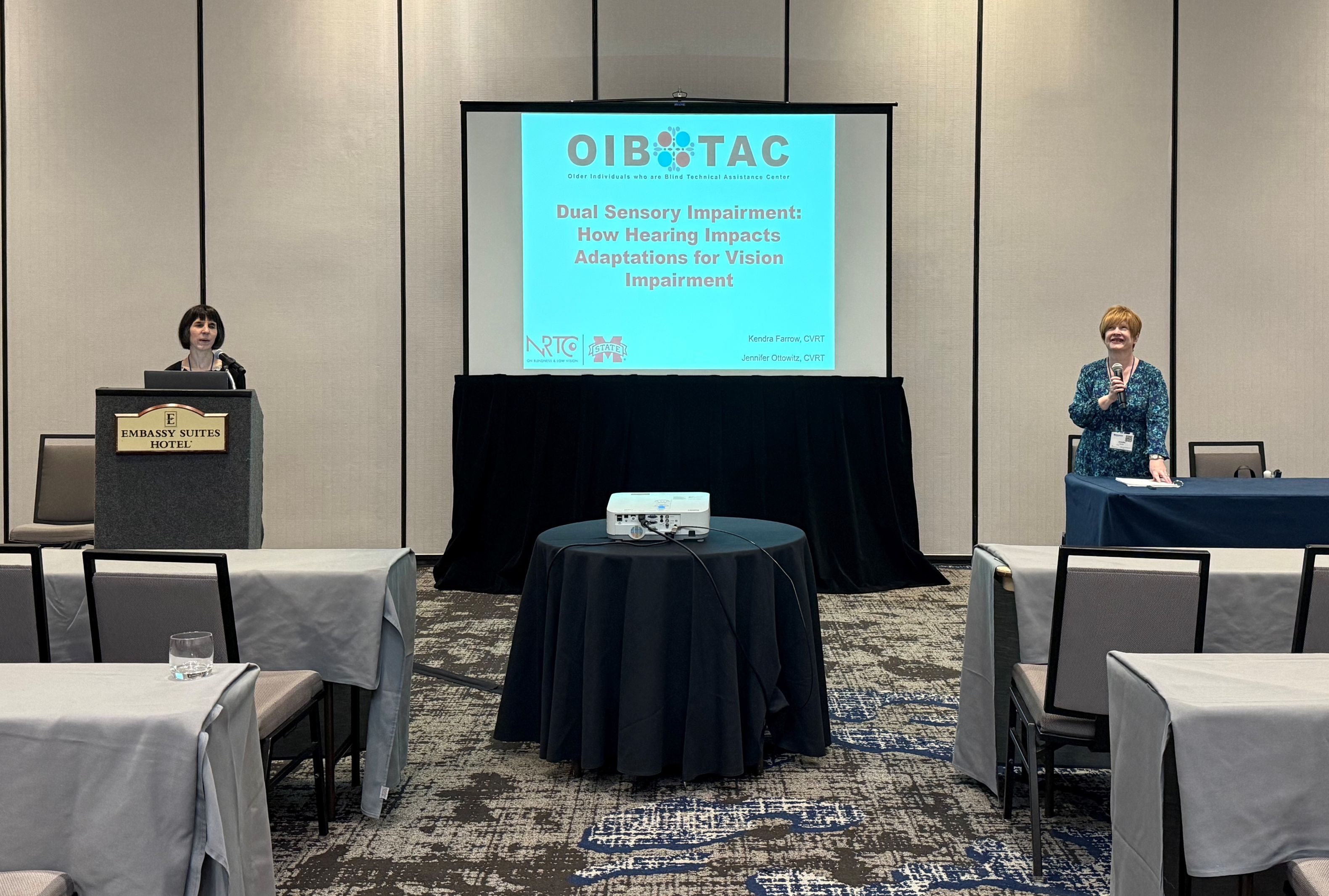 Jennifer and Kendra are standing on either side of a presentation screen at the front of a conference room. The screen displays the title “Dual Sensory Impairment: How Hearing Impacts Adaptations for Vision Impairment” with logos for OIB-TAC, NRTC, and Mississippi State University. Kendra is standing at a podium, and Jennifer is speaking from a table.