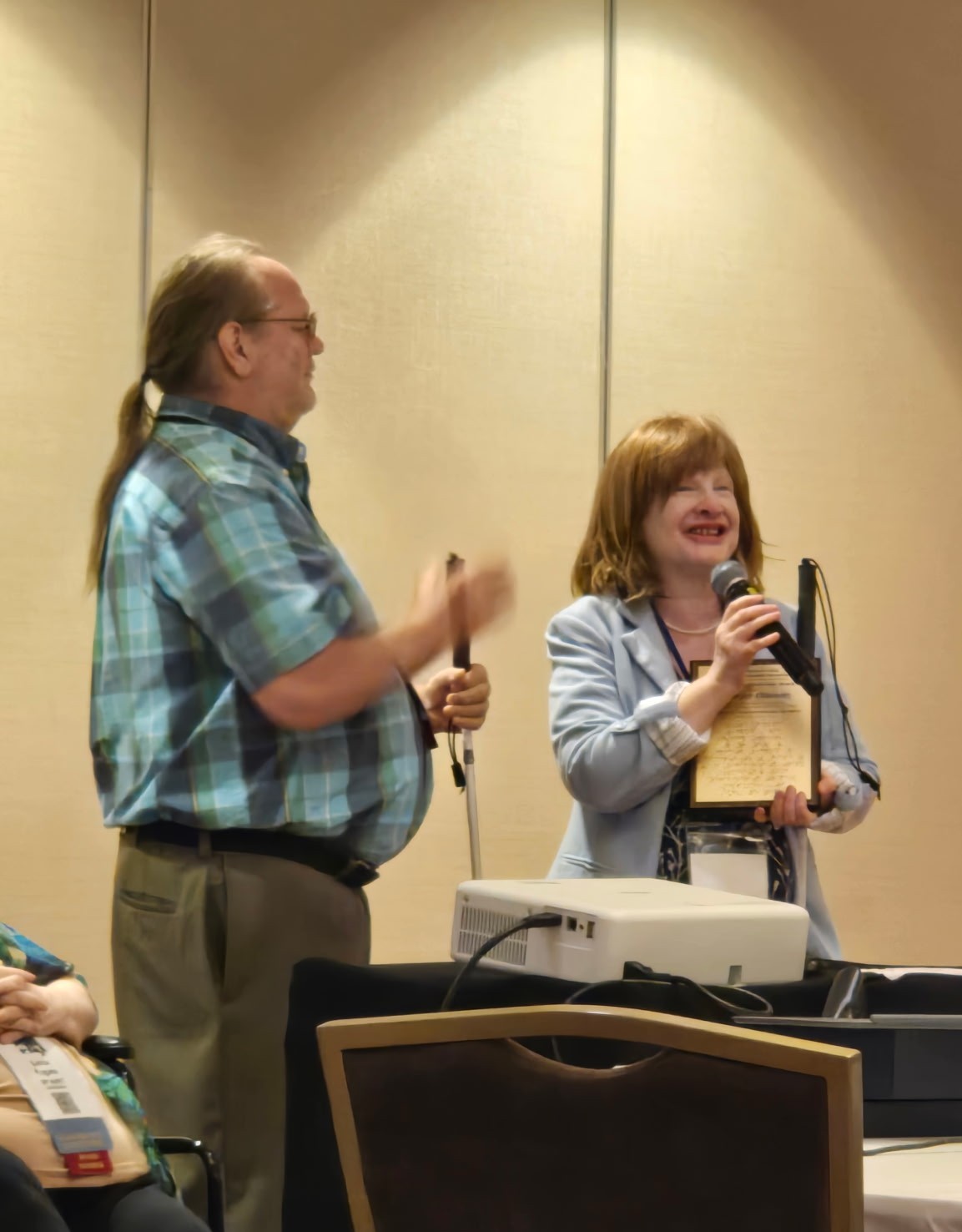 John McMahon and Jennifer stand together in a conference room. Jennifer, holding a microphone and a plaque, smiles while speaking. John stands beside her holding a white cane.