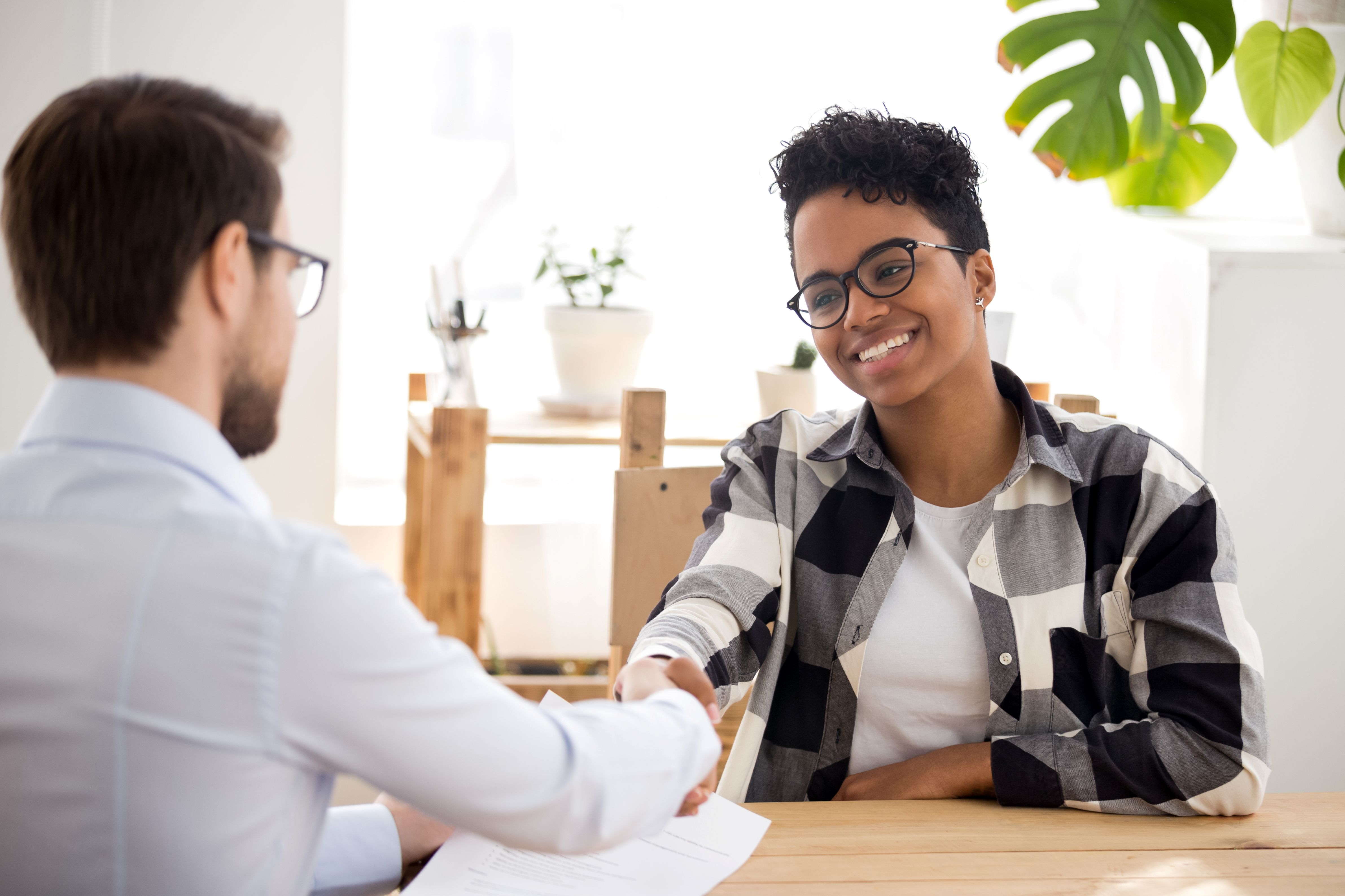 A smiling woman in a plaid shirt shakes hands with a man in an office setting, suggesting a successful job interview or agreement.