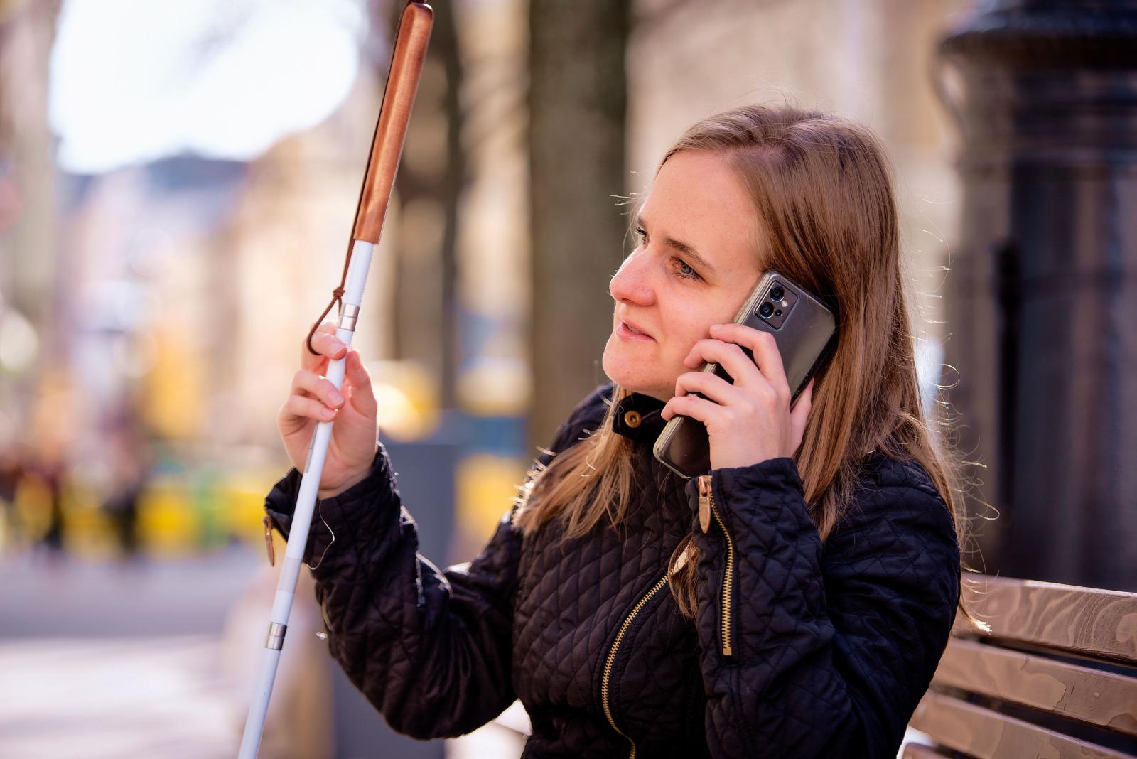 Woman seated on an outdoor bench using a smartphone while holding a white mobility cane with a brown handle. The background shows a blurred city street with trees and buildings in daylight.