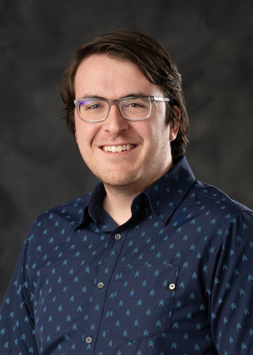 Professional headshot of Isaiah Postenrieder. Isaiah has dark brown hair and square-framed glasses, and is smiling in a navy button-up shirt.