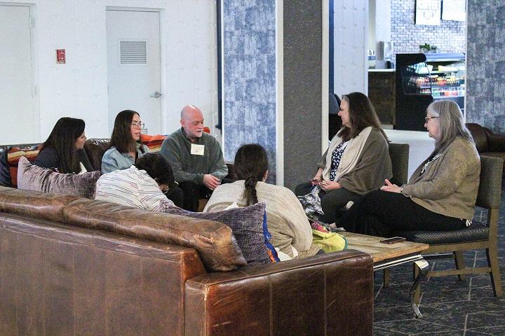 A casual meeting scene in a lounge area. A group of professionals sits on and around a large leather couch, engaged in a focused and relaxed conversation.