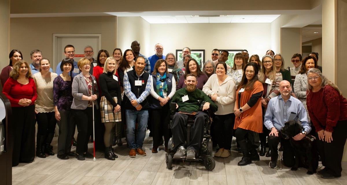 A large group of conference attendees poses for a group photo, smiling. Some individuals are using mobility aids, including a white cane and a guide dog.