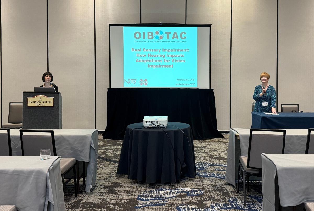 Jennifer and Kendra are standing on either side of a presentation screen at the front of a conference room. The screen displays the title “Dual Sensory Impairment: How Hearing Impacts Adaptations for Vision Impairment” with logos for OIB-TAC, NRTC, and Mississippi State University. Kendra is standing at a podium, and Jennifer is speaking from a table.