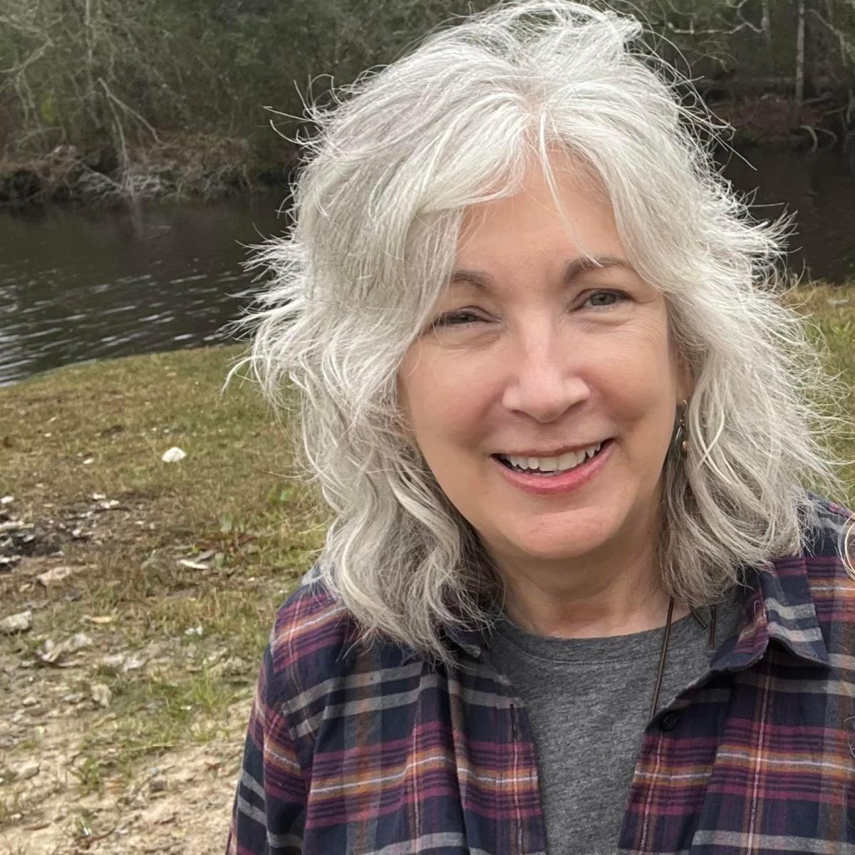 Adele Crudden smiling and wearing a plaid shirt. She standing outside by a river.